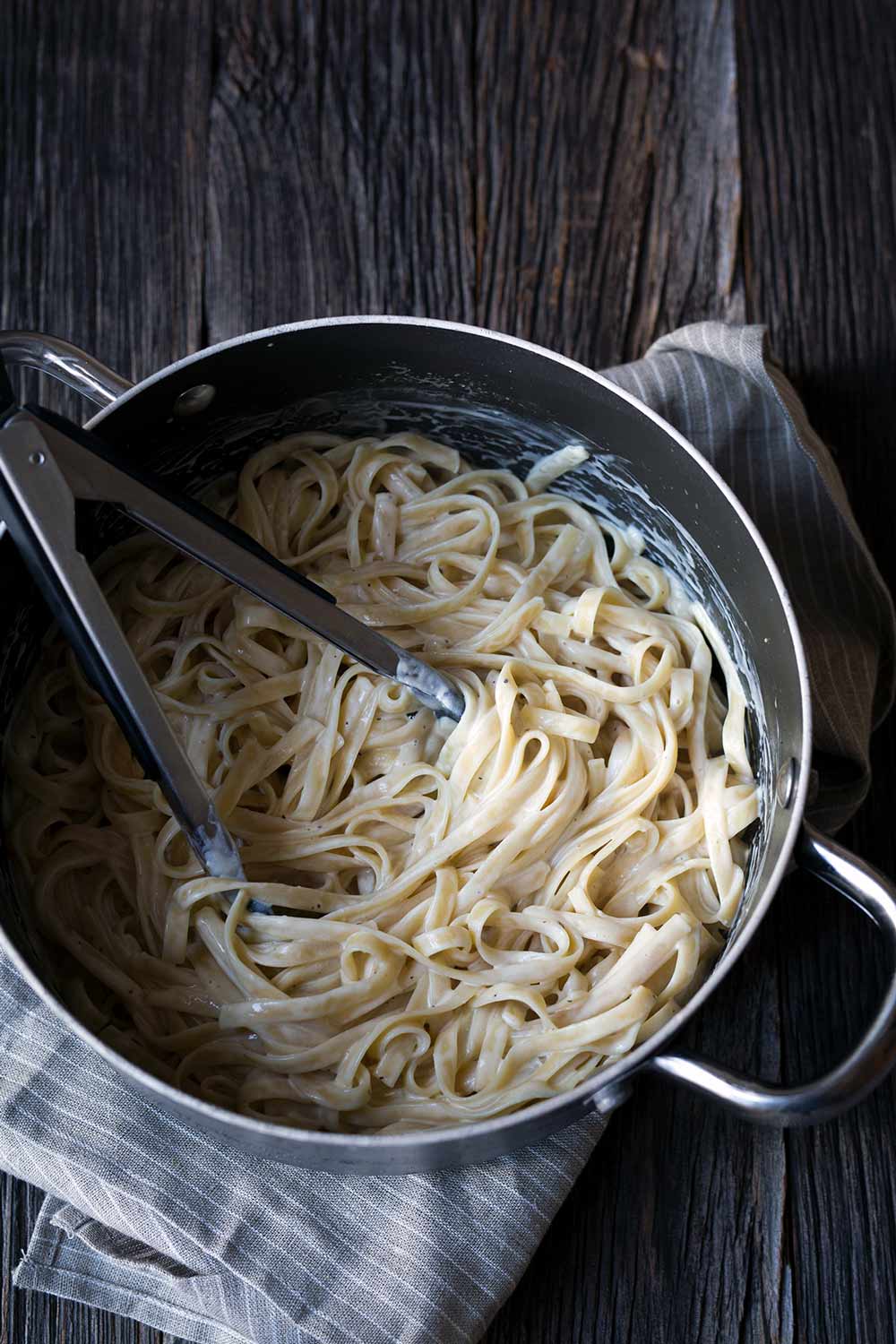A photo of vegan fettuccine alfredo in a large saucepan with tongs.