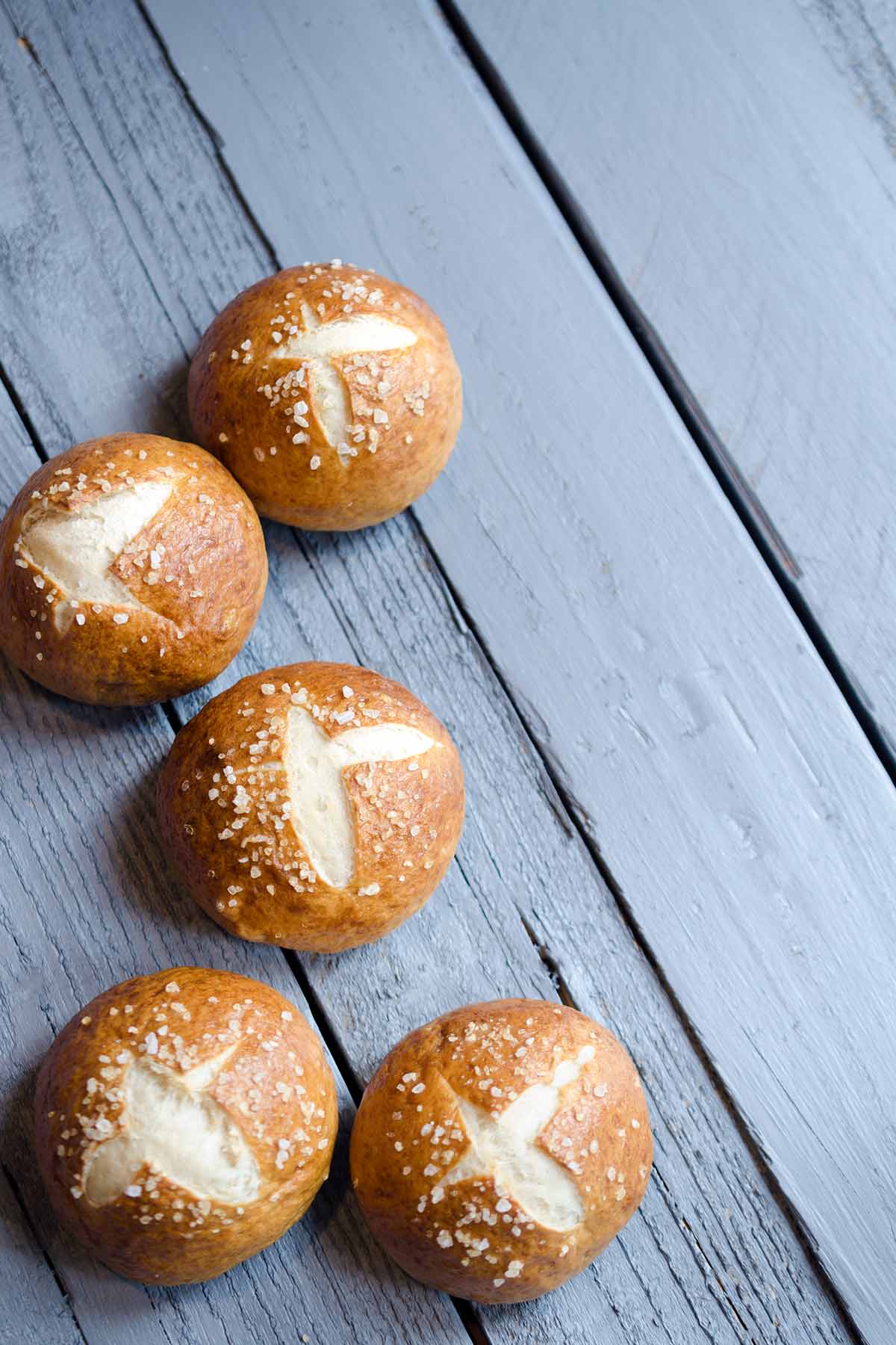 Photo of the finished pretzel bun recipe. Multiple pretzel rolls on a wood backdrop.