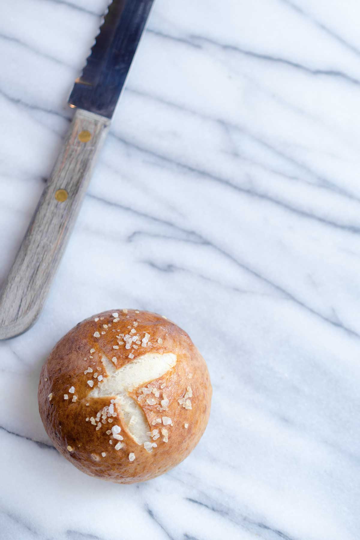 A pretzel bun on a marble backdrop next to a serrated knife.