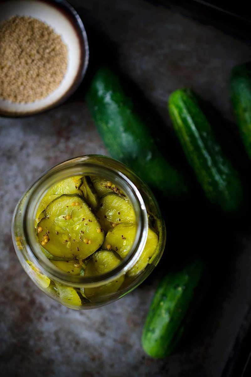 Overhead photo of bread and butter pickles recipe next to mustard seeds and cucumbers
