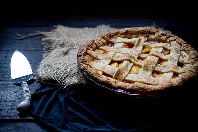 Homemade nectarine pie, uncut, on a burlap sac next to a pie cutter