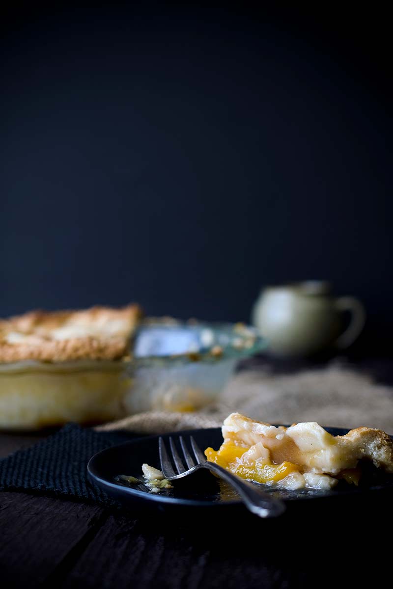 A slice of nectarine pie on a plate with a fork. Pie in dish in the background