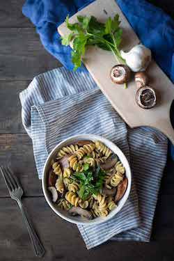 A bowl of sausage mushroom pasta on a napkin, surrounded by a fork, garlic, mushrooms, parsley, and a cutting board.