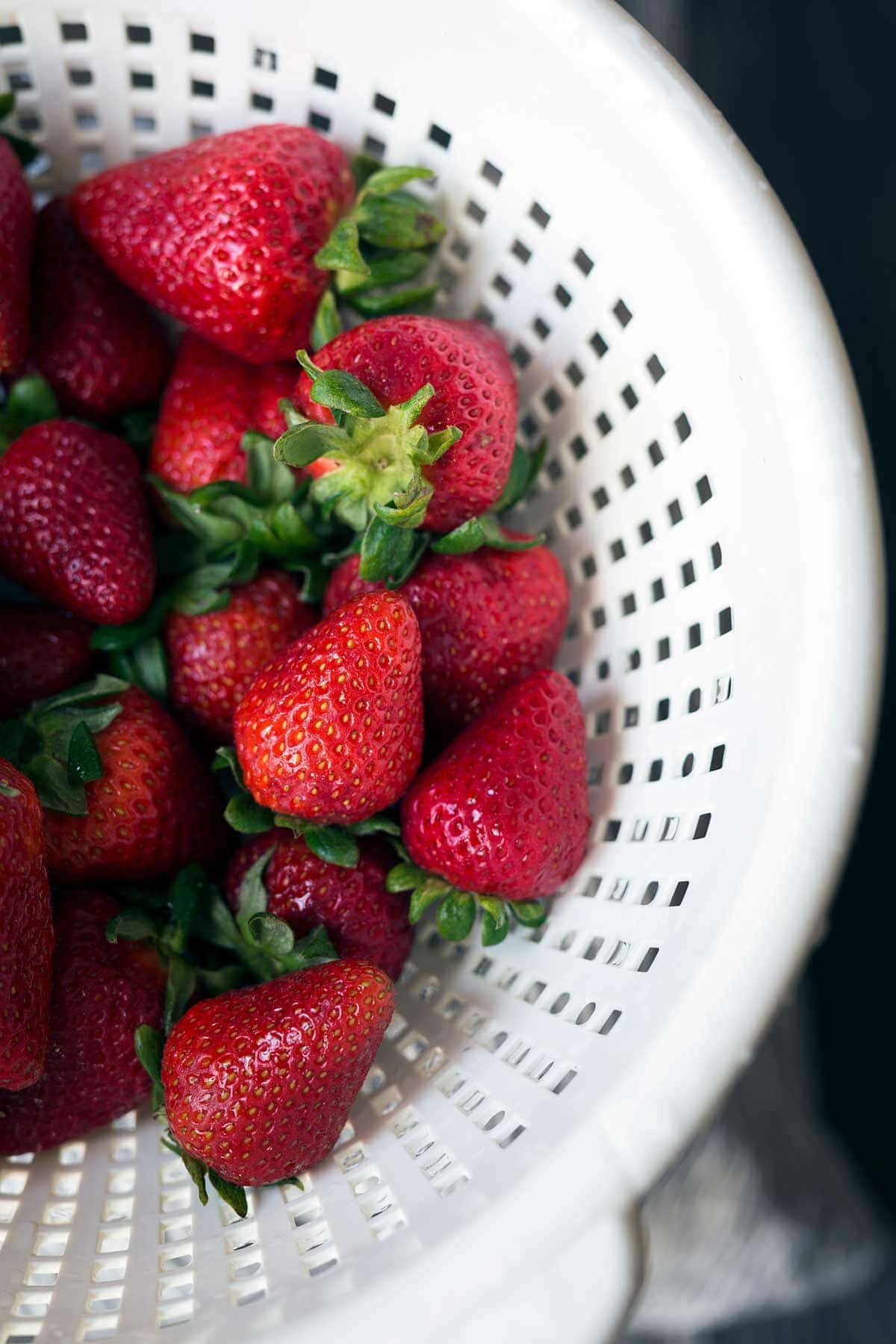Fresh strawberries with bright green leaves in a white colander.