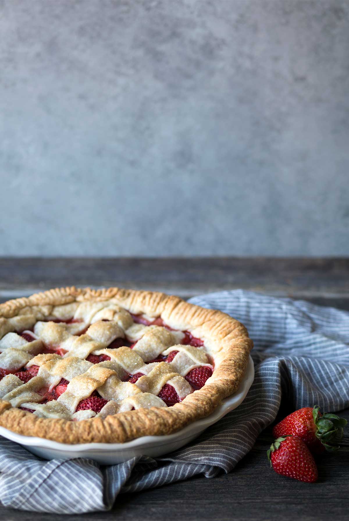 Fresh strawberry pie with a lattice top, sitting on a blue toned napkin next to fresh strawberries.