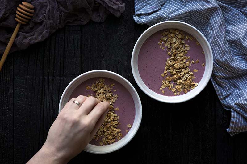 A hand sprinkling granola on top of two smoothie bowls.