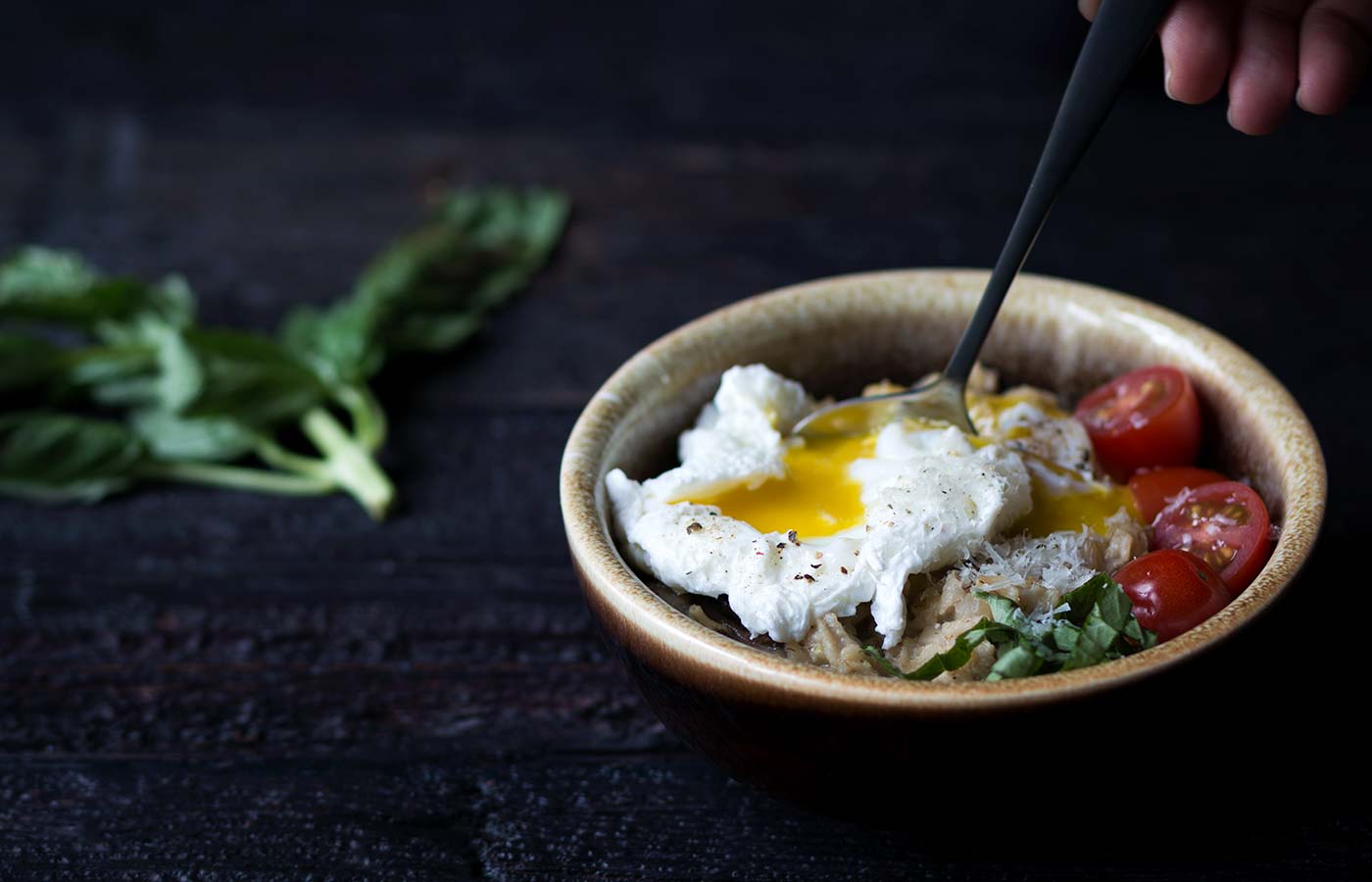 spoon breaking poached egg yolk in a bowl of cooked oats, tomatoes, and fresh basil