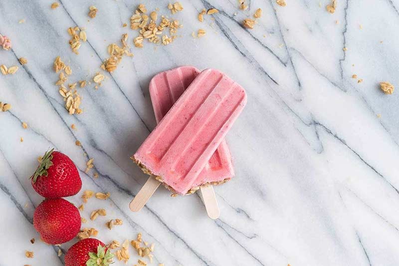 A picture of two frozen yogurt treats on wooden sticks, surrounded with whole strawberries and granola 