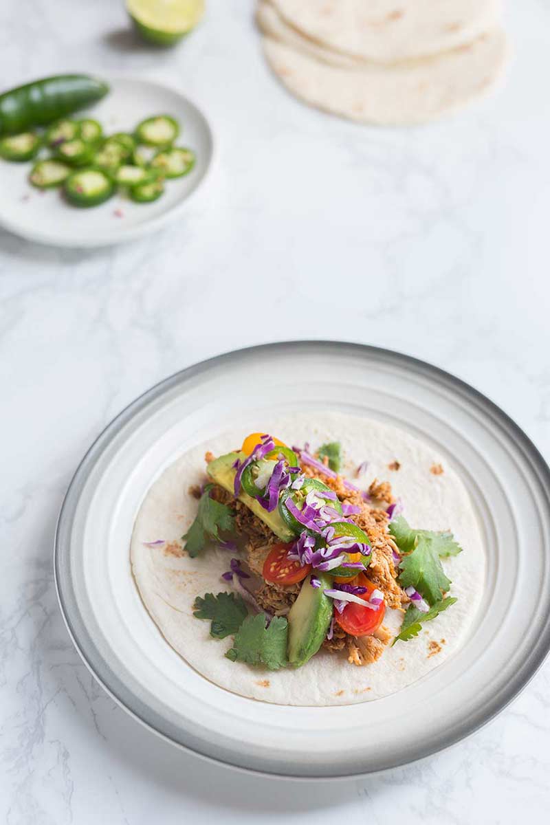 A rotisserie chicken taco recipe on a plate, surrounded by fixings. Marble backdrop. 