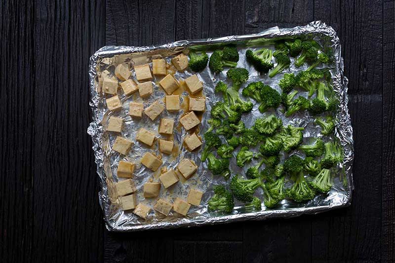 A photo of marinated tofu and broccoli on a foil-lined baking sheet, ready to be baked.