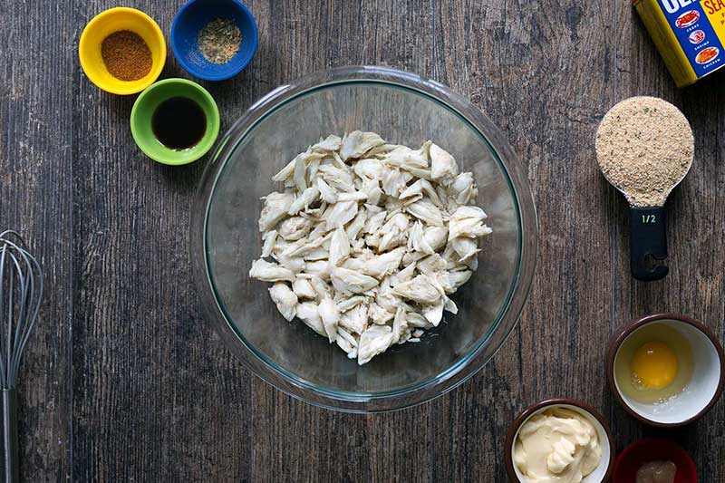 A photo of crab meat in a bowl, surrounded by the other ingredients for this baked crab cake recipe.