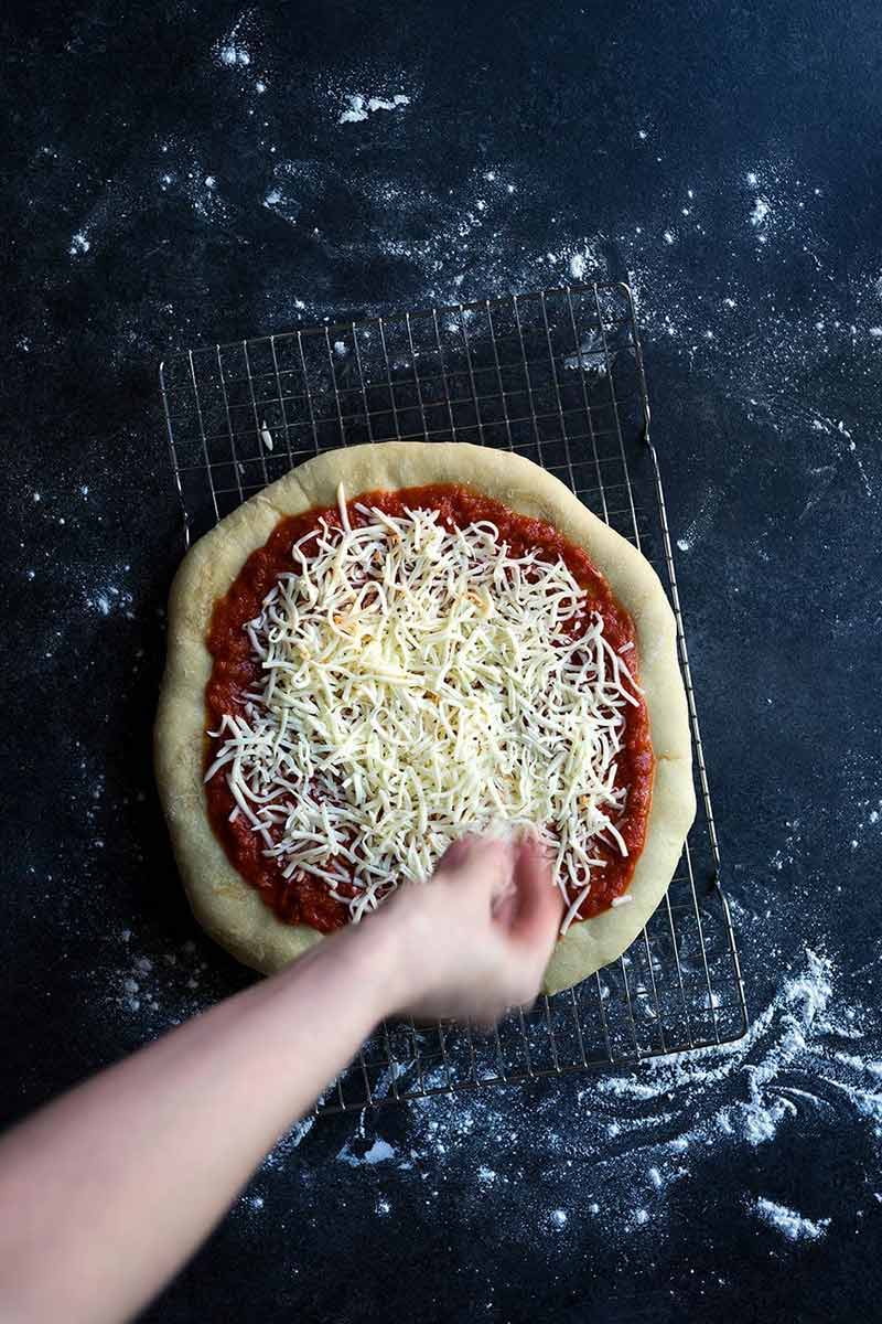 A photo of shredded mozzarella cheese being sprinkled over homemade pizza dough and sauce.