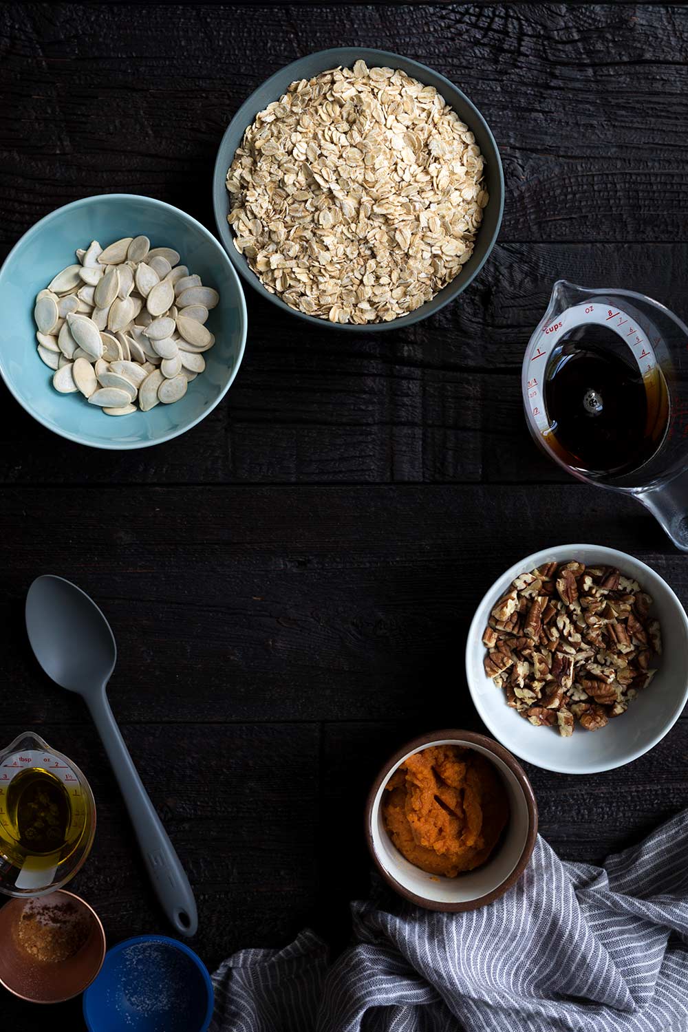 Ingredients to prepare pumpkin granola, including rolled oats, pumpkin seeds, dark amber maple syrup, pecans, pumpkin, and spices.
