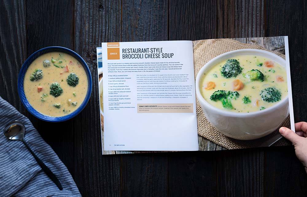 A photo of cheesy broccoli cheddar soup in a bowl next to The Simple Kitchen Cookbook, opened to the page that displays the recipe.