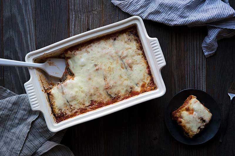 A photo of freshly baked eggplant mushroom lasagna, with one slice served up on a plate.