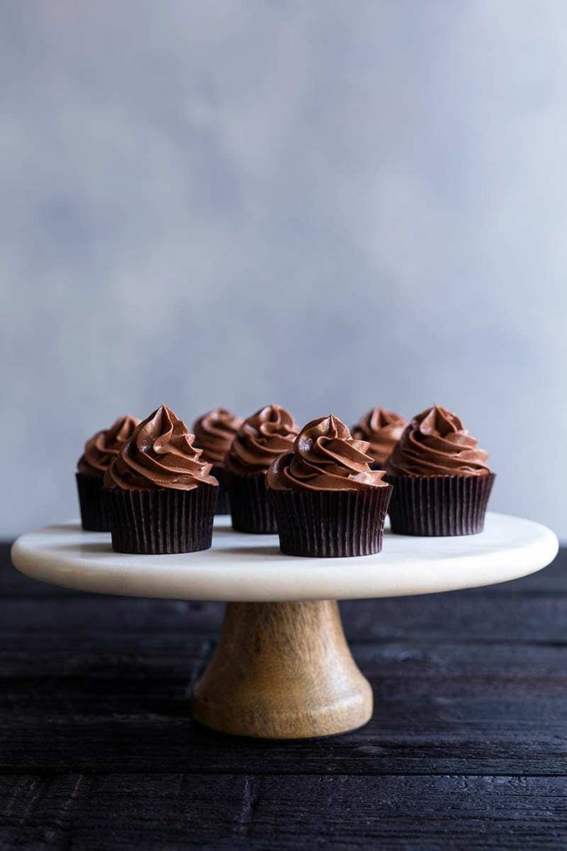 batch of homemade dark chocolate frosted cupcakes on marble and wood cake stand