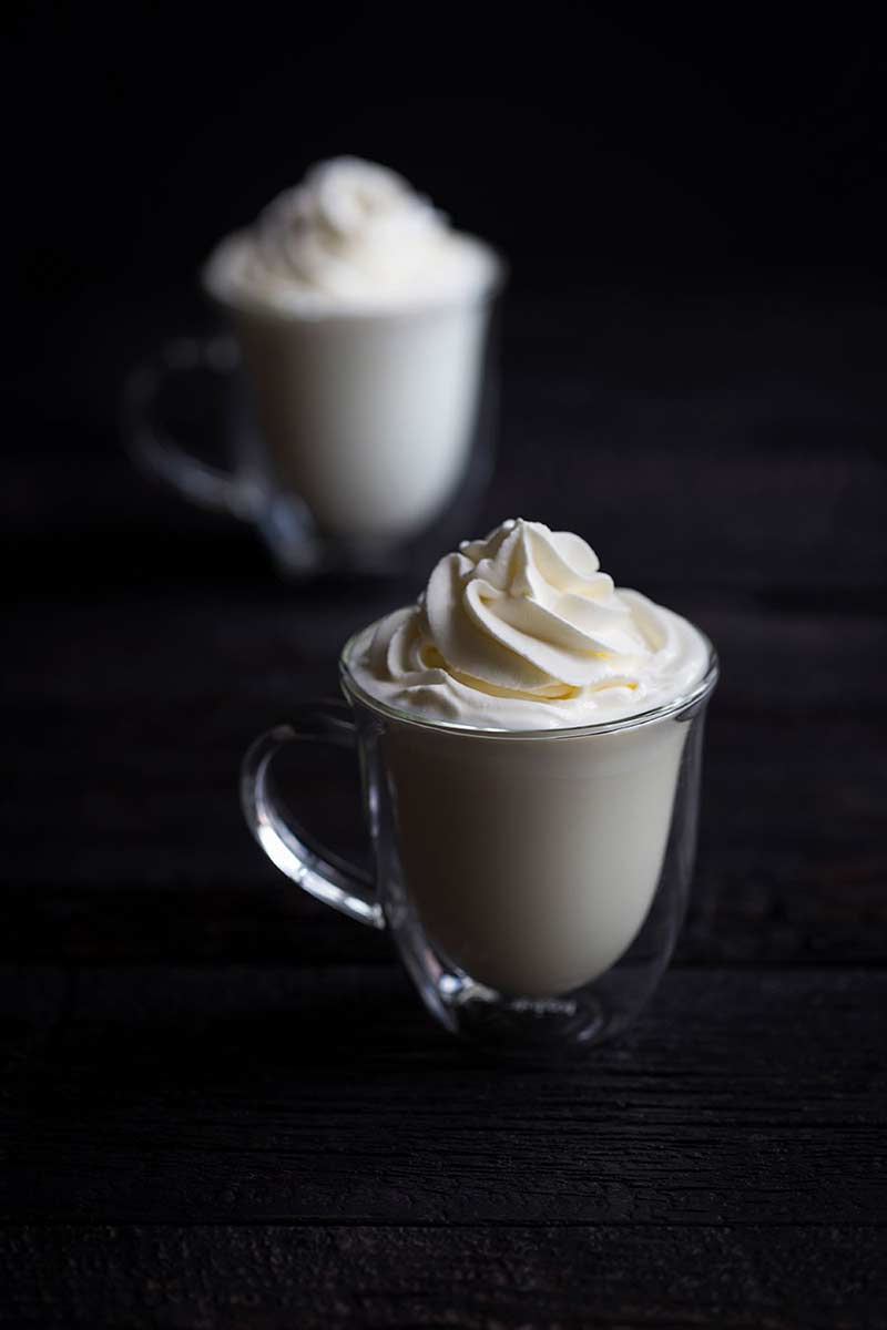 two clear glass mugs of hot white chocolate topped with whipped cream