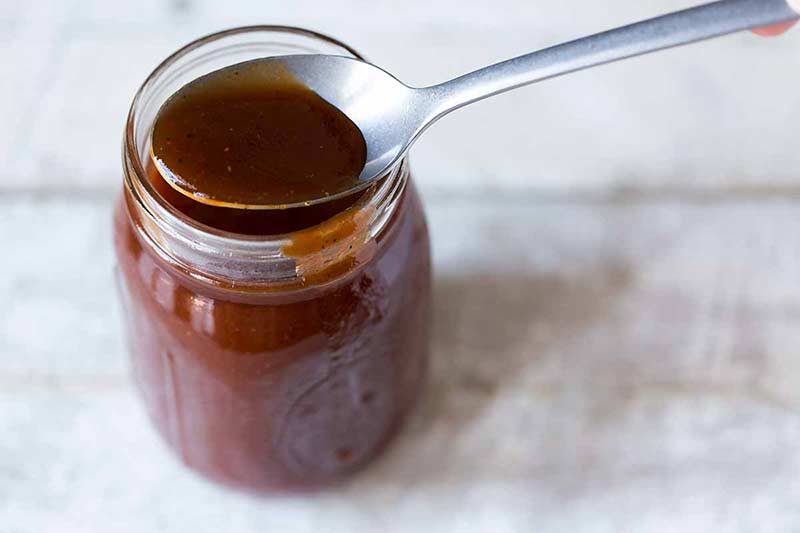 BBQ sauce recipe spilling slightly over the side of a jar, plus a spoon offering a closeup look at some of the homemade sauce.