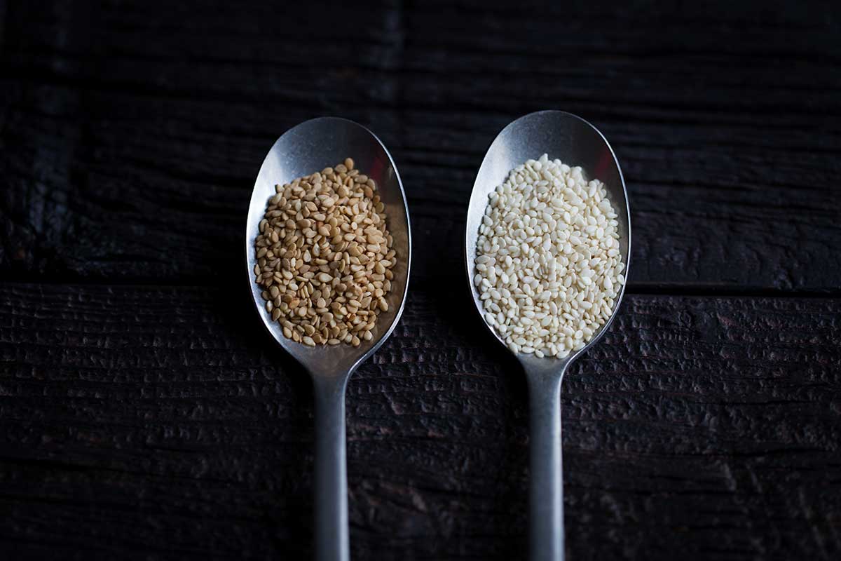 Toasted sesame seeds in a spoon next to sesame seeds that have not been toasted