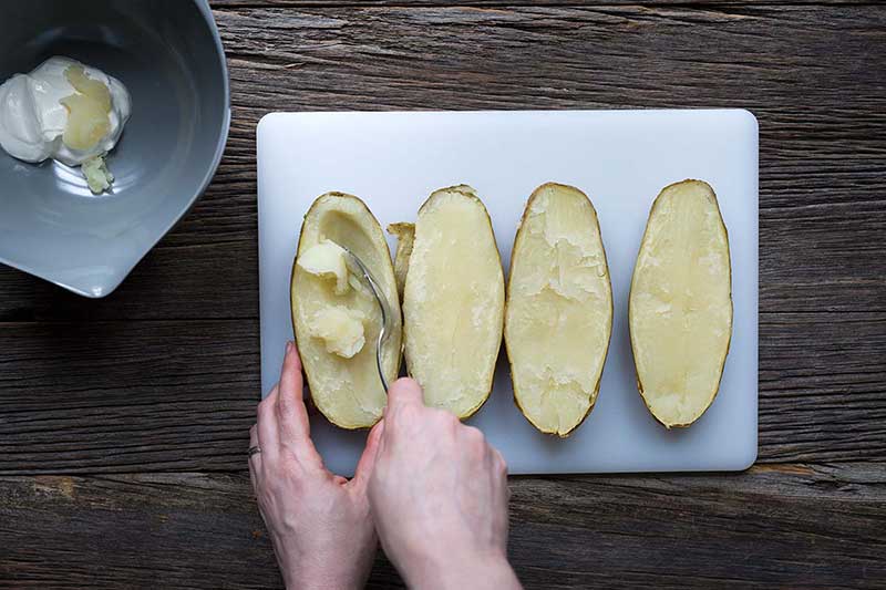 Hands scooping a the filling from a baked potato next to a large bowl with sour cream.
