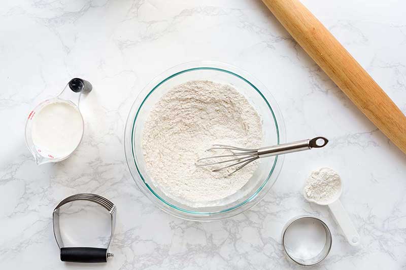 Ingredients and tools used to prepare buttermilk biscuits, including flour, buttermilk, rolling pin, whisk, and pastry blender.