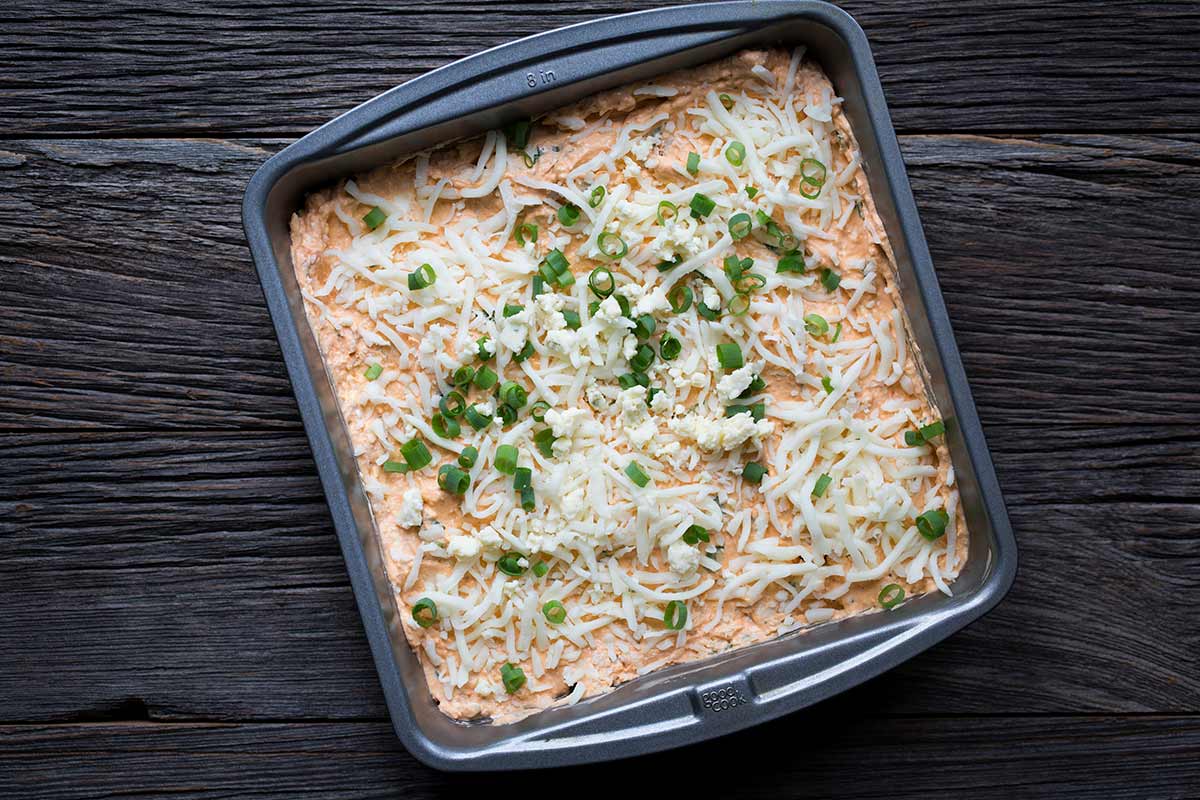 Ingredients for buffalo chicken dip, layered in an 8x8 pan, ready to bake.
