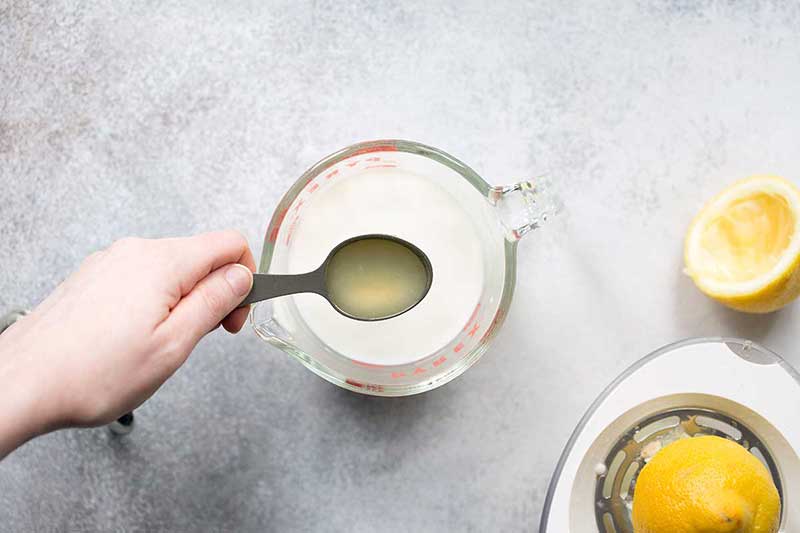 A hand holding a tablespoon of lemon juice over a cup of milk.