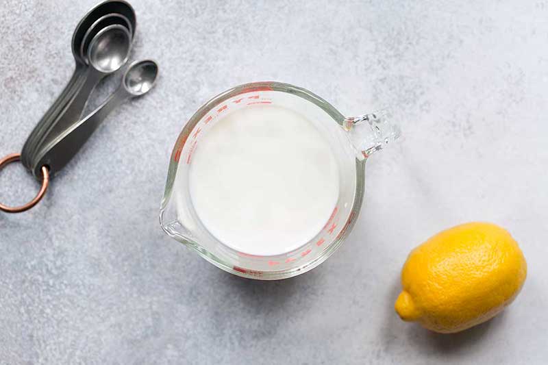 Milk in a pyrex measuring cup, next to a lemon and measuring spoons.