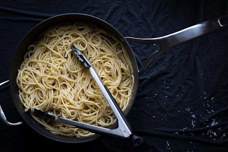 A photo of Cacio e Pepe recipe immediately after cooking, in a flat-sided skillet with tongs sitting on top..