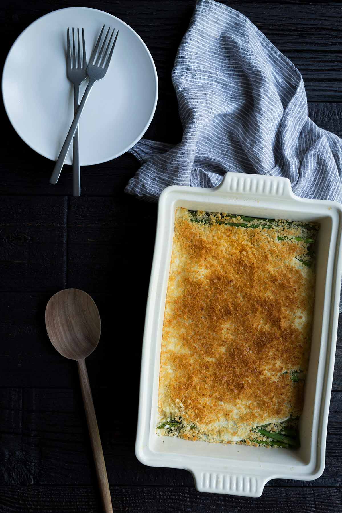 Baked asparagus gratin surrounded by a plate, forks, a serving spoon, and a napkin.