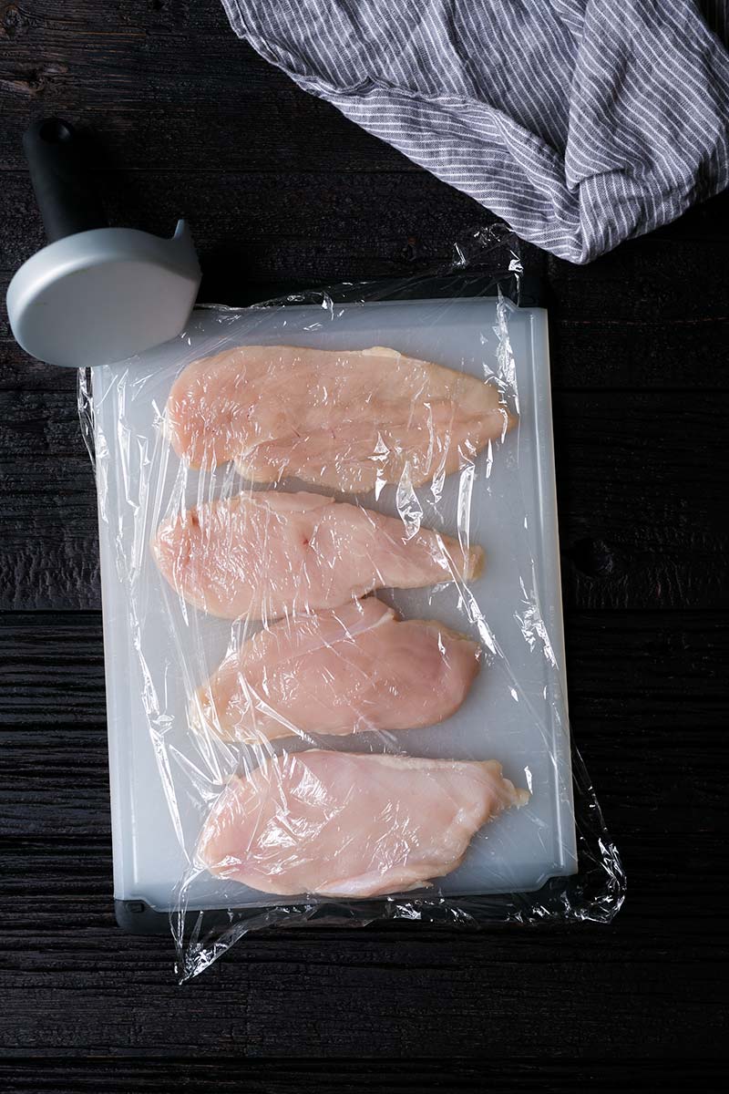 Raw, butterflied chicken breasts on a cutting board, topped with plastic wrap, shown next to a meat tenderizer. 