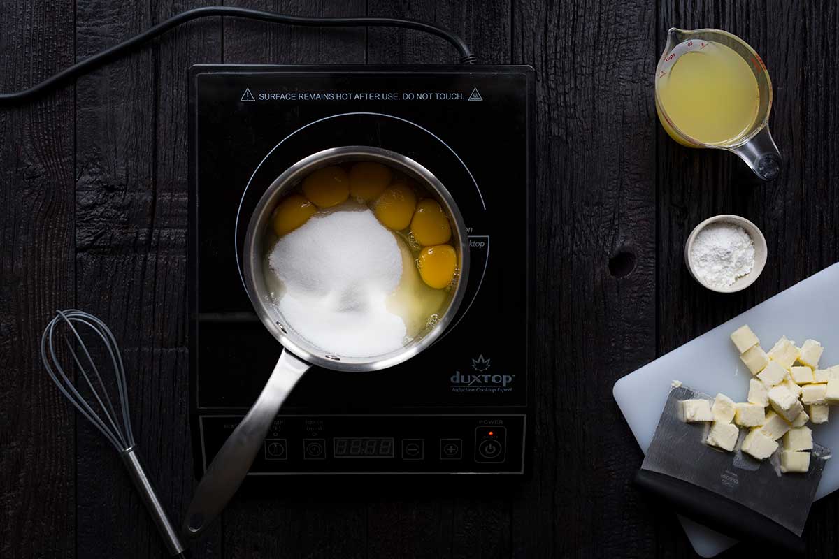 An induction burner topped with a saucepan filled with eggs and sugar, surrounded by butter, lemon juice and cornstarch.