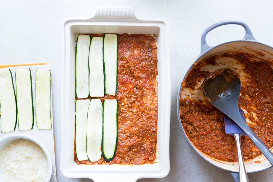 zucchini noodle lasagna being prepared, with thin layers of zucchini spread on top of a layer of sauce.
