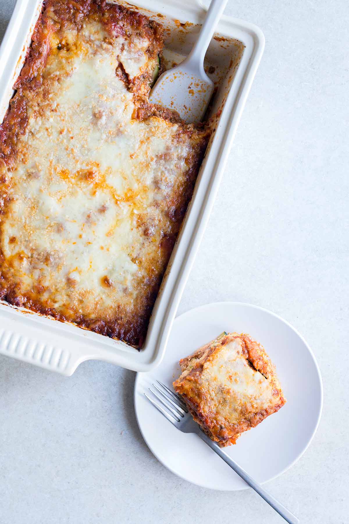 zucchini lasagna recipe in a casserole dish, next to a slice of lasagna on a plate.