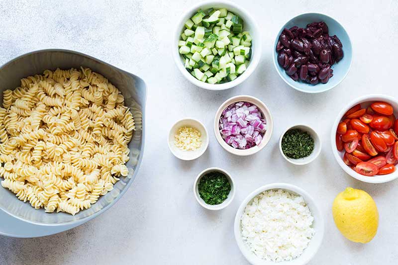 summer salad recipe ingredients, including cooked pasta, cucumbers, olives, tomatoes, red onions, garlic, and feta cheese.