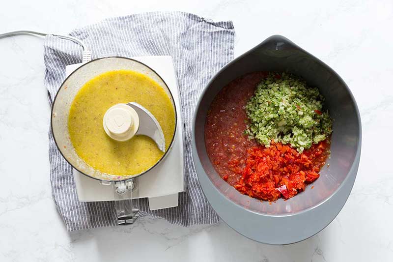 A large bowl full of assorted gazpacho ingredients next to a food processor filled with pureed olive oil, red onions and garlic.