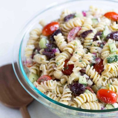 A close up shot of Greek pasta salad in a bowl, next to a wood spoon.