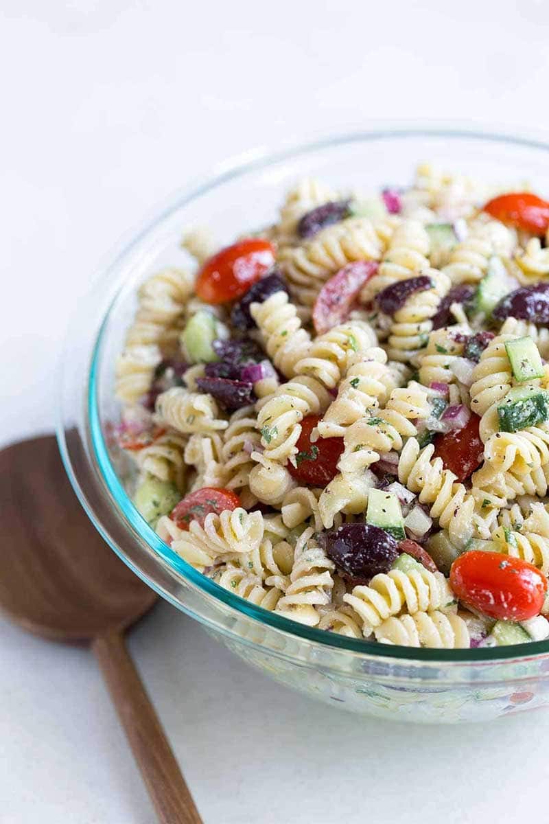 A close up shot of Greek pasta salad in a bowl, next to a wood spoon.