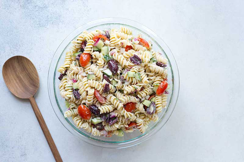 Summer rotini pasta salad in a bowl on a pale backdrop next to a wood spoon.