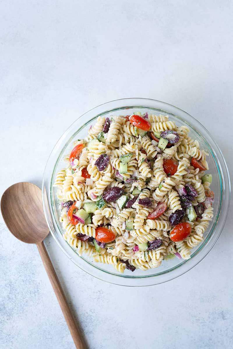 overhead image: large wooden serving spoon next to bowl of Greek pasta salad