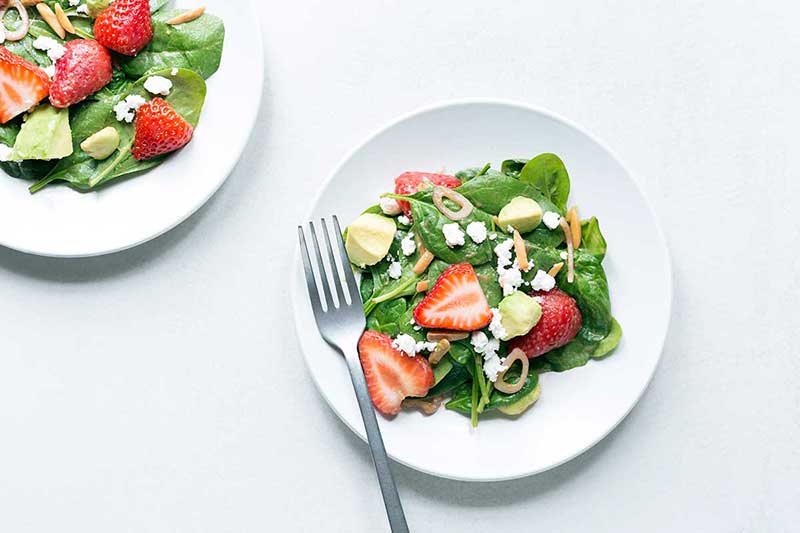 Photo of two spinach salads topped with strawberries, avocados, feta, almonds, and shallots. Shot on a white backdrop.