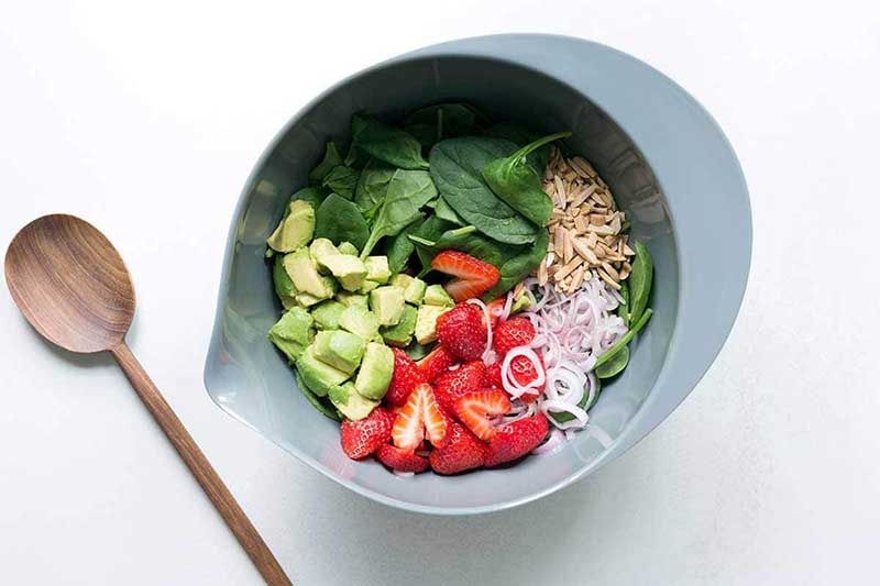 A grey bowl filled with ingredients for spinach strawberry salad, next to a wood spoon.