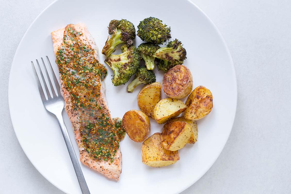 Baked salmon with potatoes and broccoli on a white plate, topped with a sauce made from whole grain mustard and chives.