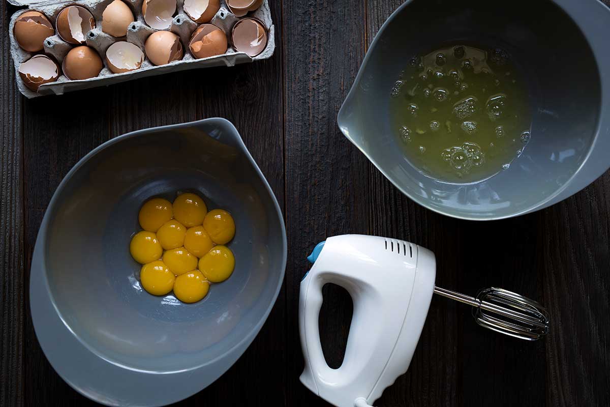 Egg whites and egg yolks separates into separate bowls, next to an electric hand mixer.