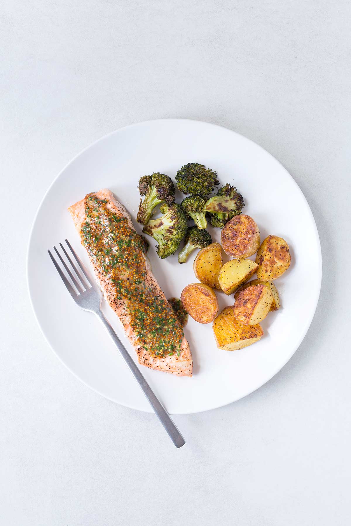 Roasted salmon, potatoes, and broccoli on a white plate with a fork, topped with a sauce.