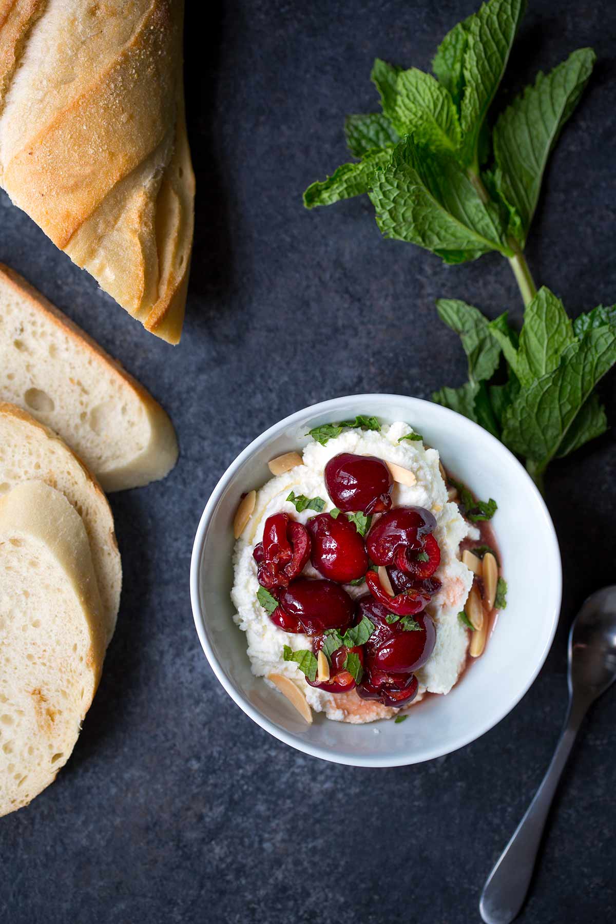 A photo of cherries macerated in honey and sherry vinegar, served over ricotta cheese and topped with almonds and mint. 