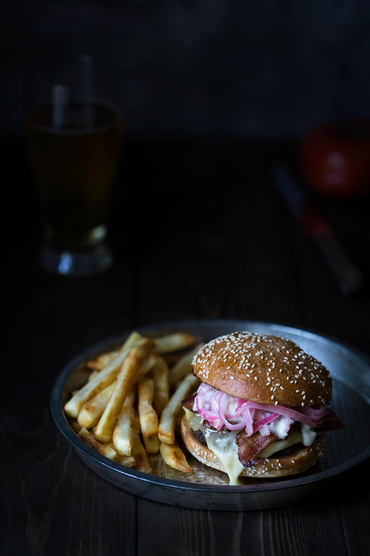 A raclette bacon cheeseburger topped with pickled shallots, horseradish aioli, served on a plate with french fries.