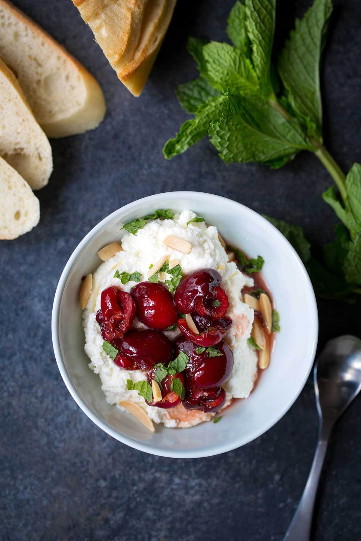 Macerated Cherries with Ricotta, Mint, and Almonds, served in a bowl. Surrounded by crostini and fresh mint on a blue backdrop.
