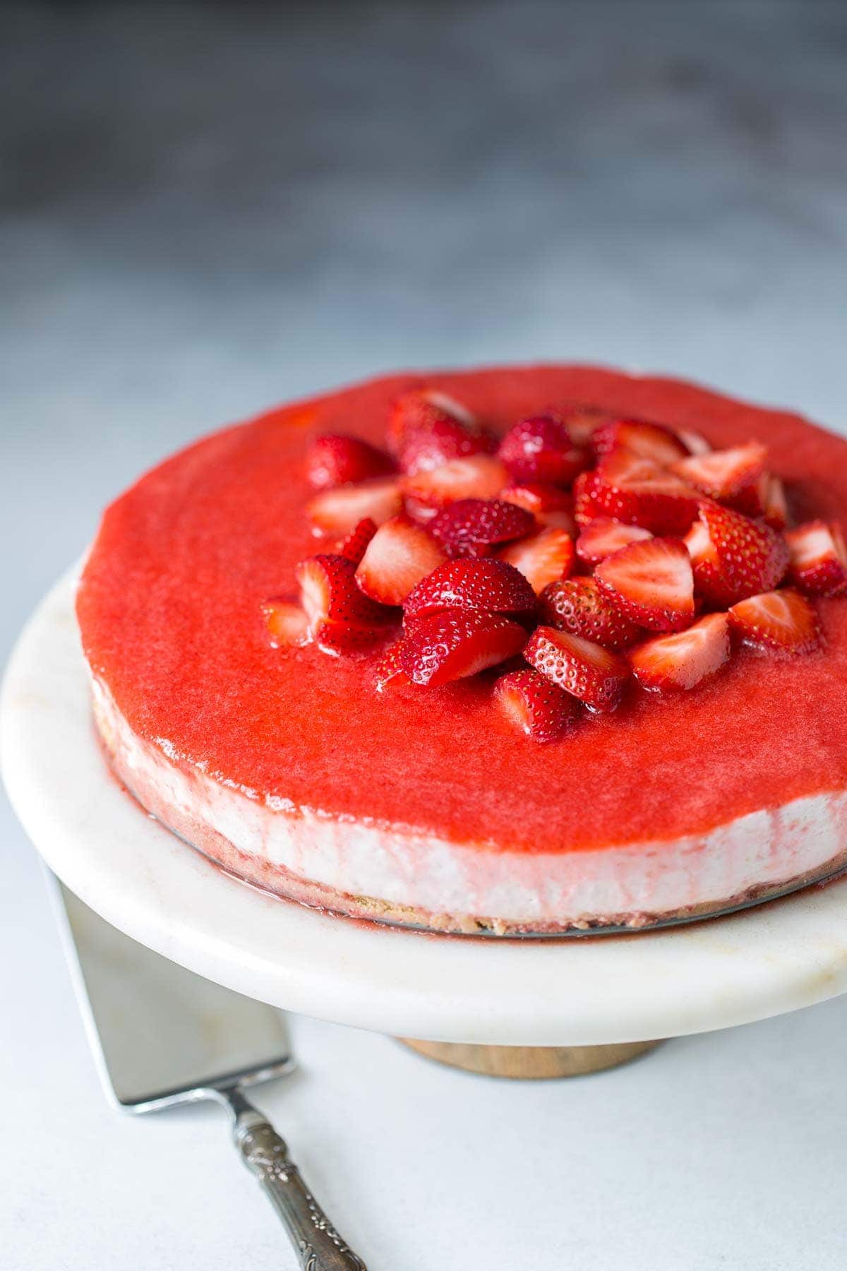 Strawberry No-Bake Cheesecake showcased on a marble and wood cake stand with a silver cake slicer. 