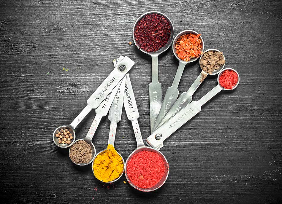 Two sets of measuring spoons filled with various colorful spices, set on a dark backdrop. Learn how many teaspoons are in a tablespoon.
